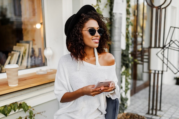 Joven mujer mixta con peinado afro hablando por teléfono móvil y sonriendo en segundo plano urbano. Chica negra con ropa casual. Sosteniendo la taza de café. Sombrero negro.