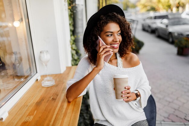 Joven mujer mixta con peinado afro hablando por teléfono móvil y sonriendo en segundo plano urbano. Chica negra con ropa casual. Sosteniendo la taza de café. Sombrero negro.