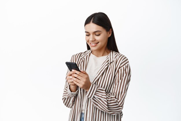 Joven mujer moderna con un atuendo elegante usando su teléfono móvil chateando en una aplicación de teléfono inteligente y una pantalla de lectura sonriente de pie sobre fondo blanco