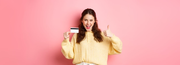 Foto gratuita joven mujer moderna diciendo sí y mostrando los pulgares hacia arriba sosteniendo una tarjeta de crédito de plástico de pie sobre rosa b