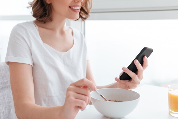 joven mujer morena en camiseta blanca con smartphone mientras desayuna con copos de maíz