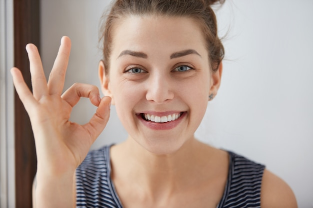 Joven mujer morena europea mostrando OK-gesto con sus dedos. Mujer feliz en top rayado sonriendo con ojos azules. Su boca de dientes blancos y su cara feliz demuestran que todo va según el plan.