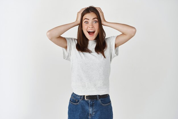 Joven mujer morena mirando sorprendido al frente, grito de alegría y emoción, tomados de la mano en la cabeza por la incredulidad, de pie sobre la pared blanca