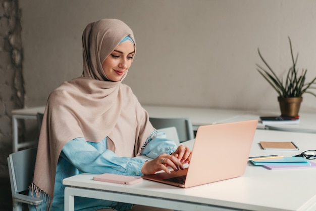 Joven mujer musulmana bastante moderna en hijab trabajando en la computadora portátil en la sala de oficina, educación en línea