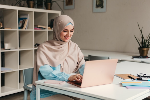 Joven mujer musulmana bastante moderna en hijab trabajando en la computadora portátil en la sala de oficina, educación en línea