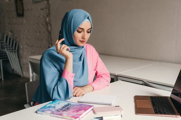 Joven mujer musulmana bastante moderna en hijab trabajando en la computadora portátil en la sala de oficina, educación en línea