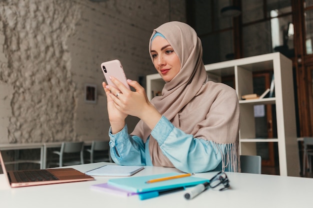 Joven mujer musulmana bastante moderna en hijab trabajando en la computadora portátil en la sala de oficina, educación en línea