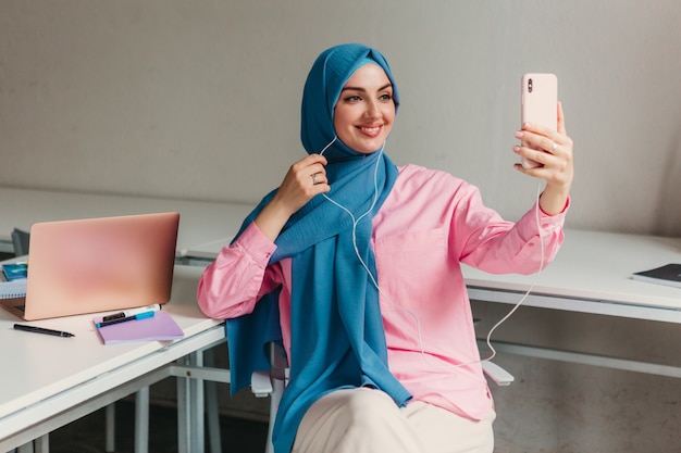 Joven mujer musulmana bastante moderna en hijab trabajando en la computadora portátil en la sala de oficina, educación en línea