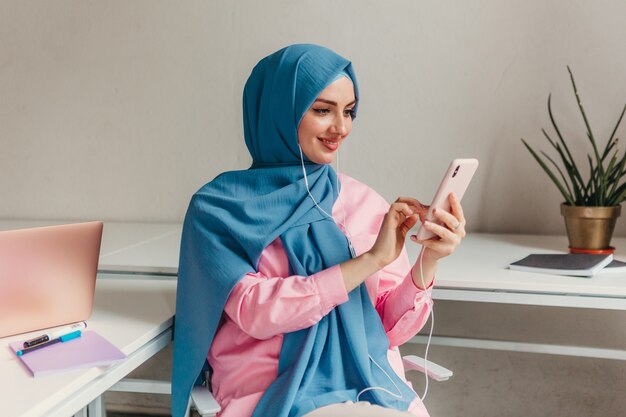 Joven mujer musulmana bastante moderna en hijab trabajando en la computadora portátil en la sala de oficina, educación en línea