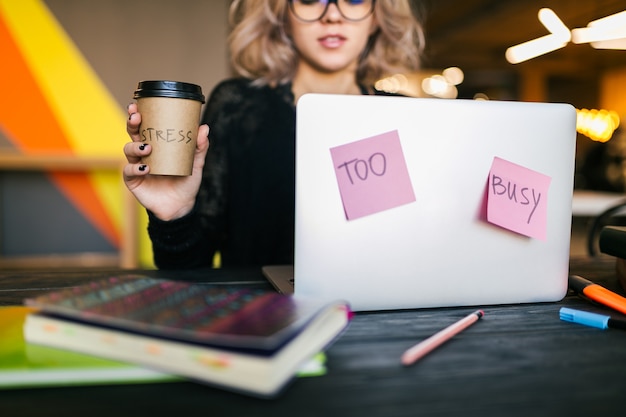 Joven mujer muy ocupada sentada en la mesa trabajando en la computadora portátil en la oficina de co-trabajo, vista cercana, consentimiento, cansado, sosteniendo la taza de café