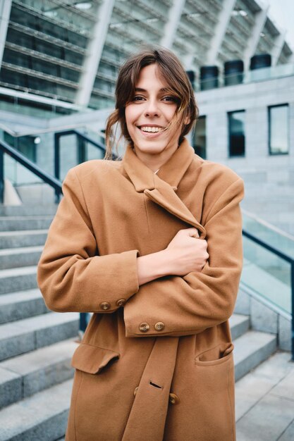 Joven mujer muy sonriente sujetando el abrigo del frío felizmente mirando a la cámara al aire libre