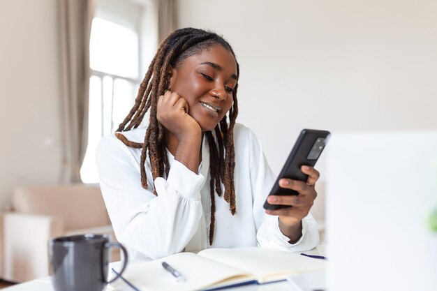 Joven mujer de negocios africana sonriente usando un teléfono inteligente cerca de la computadora en la oficina