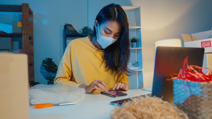 Foto gratuito joven mujer de negocios asiática con máscara facial comprobando la orden de compra en su teléfono inteligente