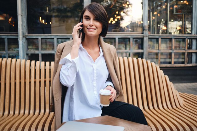 Joven mujer de negocios bastante elegante con una computadora portátil felizmente hablando por teléfono celular durante el descanso para tomar café en un café en la calle