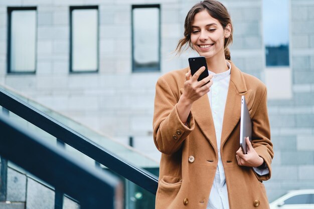 Joven mujer de negocios bastante informal con abrigo y portátil usando alegremente el teléfono móvil al aire libre