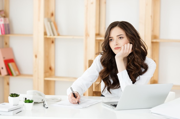 Joven mujer de negocios bonita con cuaderno y documento en la oficina moderna brillante en el interior