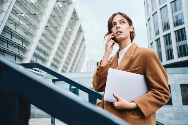 Joven mujer de negocios casual pensativa en abrigo con laptop hablando por teléfono celular mirando cuidadosamente al aire libre