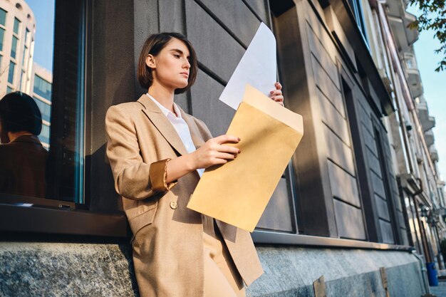 Joven mujer de negocios elegante y confiada en traje trabajando cuidadosamente con papeles en la calle de la ciudad