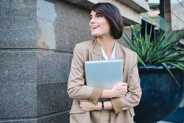 Joven mujer de negocios elegante y muy sonriente con una laptop mirando alegremente a un lado en la calle de la ciudad