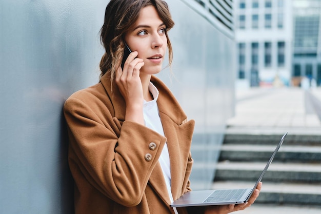 Joven mujer de negocios con estilo en abrigo hablando cuidadosamente por teléfono celular trabajando en una computadora portátil en la calle de la ciudad