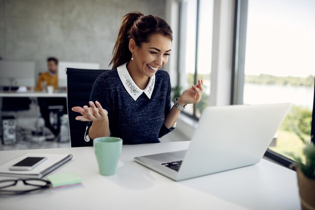 Joven mujer de negocios feliz hablando mientras hace una videollamada a través de una laptop en la oficina