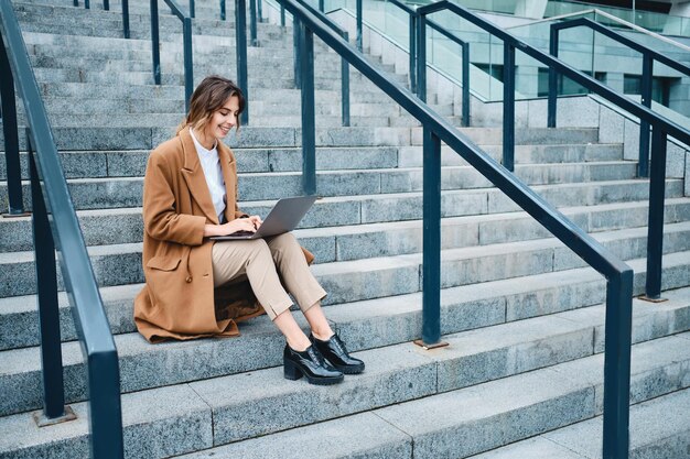 Joven mujer de negocios muy sonriente con abrigo trabajando felizmente en una laptop en las escaleras al aire libre