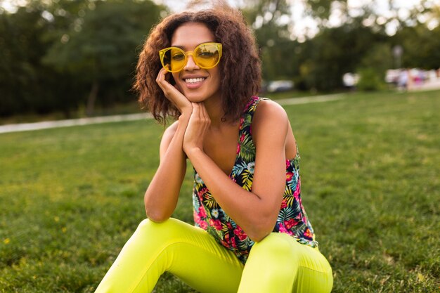 Joven mujer negra elegante divirtiéndose en el estilo de la moda de verano del parque, colorido traje hipster, sentado en la hierba con gafas de sol amarillas