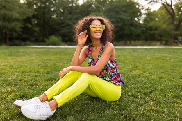 Joven mujer negra elegante divirtiéndose en el estilo de la moda de verano del parque, colorido traje hipster, sentado en la hierba con pantalones y gafas de sol amarillas, zapatillas de deporte