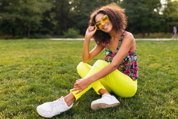 Joven mujer negra elegante divirtiéndose en el estilo de la moda de verano del parque, colorido traje hipster, sentado en la hierba con pantalones y gafas de sol amarillas, zapatillas de deporte