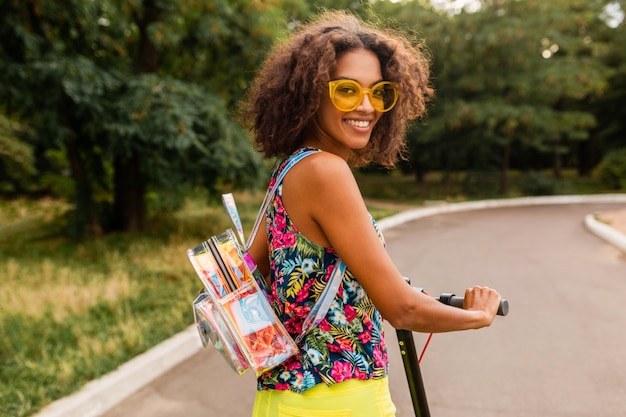 Joven mujer negra elegante divirtiéndose en el parque montando en patinete eléctrico en estilo de moda de verano, colorido traje hipster, con mochila y gafas de sol amarillas