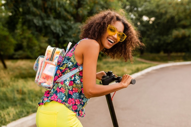 Joven mujer negra elegante divirtiéndose en el parque montando en patinete eléctrico en estilo de moda de verano, colorido traje hipster, con mochila y gafas de sol amarillas