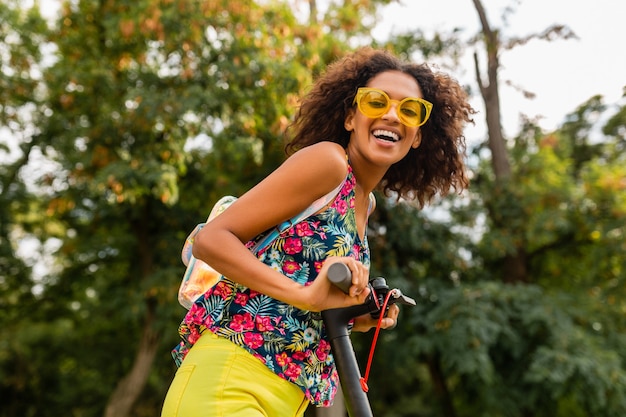 Joven mujer negra elegante divirtiéndose en el parque montando en patinete eléctrico en estilo de moda de verano, colorido traje hipster, con mochila y gafas de sol amarillas