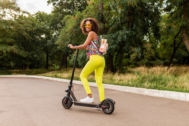Joven mujer negra elegante divirtiéndose en el parque montando en patinete eléctrico en estilo de moda de verano, colorido traje hipster, con mochila y pantalones amarillos y gafas de sol