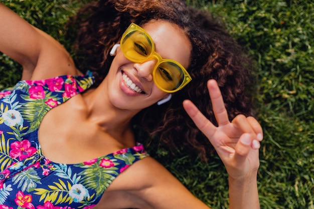 Joven mujer negra con estilo escuchando música con auriculares inalámbricos divirtiéndose en el parque, estilo de moda de verano, traje de hipster colorido, tumbado en la hierba, vista desde arriba