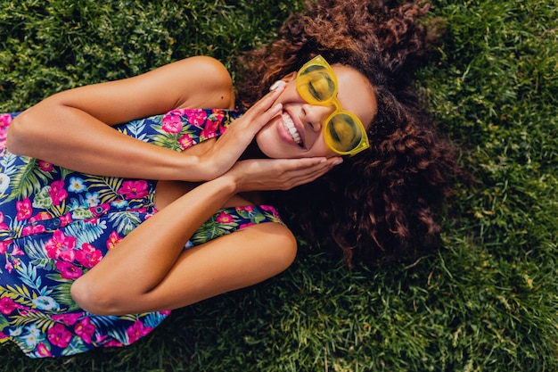 Joven mujer negra con estilo escuchando música con auriculares inalámbricos divirtiéndose en el parque, estilo de moda de verano, traje de hipster colorido, tumbado en la hierba, vista desde arriba