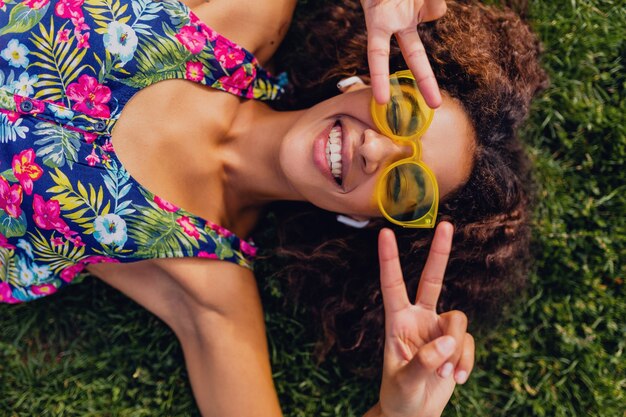 Joven mujer negra con estilo escuchando música con auriculares inalámbricos divirtiéndose tumbado en la hierba en el parque, estilo de moda de verano, traje de hipster colorido, vista desde arriba