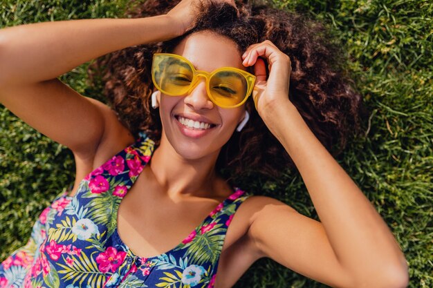 Joven mujer negra con estilo escuchando música con auriculares inalámbricos divirtiéndose tumbado en la hierba en el parque, estilo de moda de verano, traje de hipster colorido, vista desde arriba