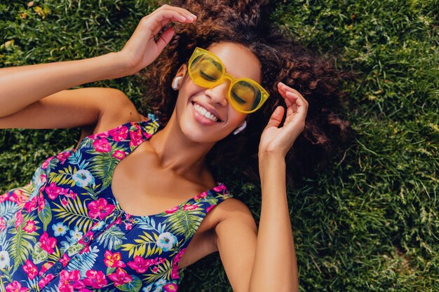 Joven mujer negra con estilo escuchando música con auriculares inalámbricos divirtiéndose tumbado en la hierba en el parque, estilo de moda de verano, traje de hipster colorido, vista desde arriba