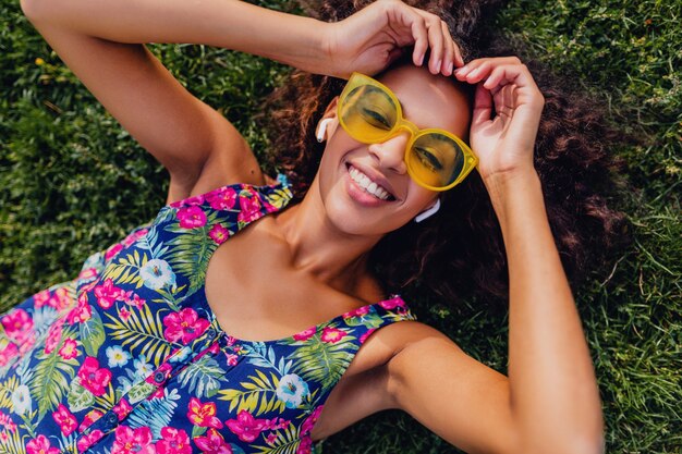 Joven mujer negra con estilo escuchando música con auriculares inalámbricos divirtiéndose tumbado en la hierba en el parque, estilo de moda de verano, traje de hipster colorido, vista desde arriba
