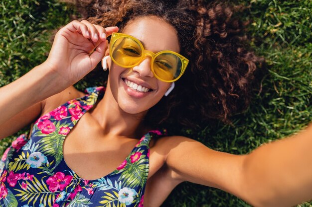 Joven mujer negra con estilo escuchando música con auriculares inalámbricos divirtiéndose tumbado en la hierba en el parque tomando foto selfie en la cámara del teléfono, traje de hipster colorido de estilo de moda de verano, vista desde arriba