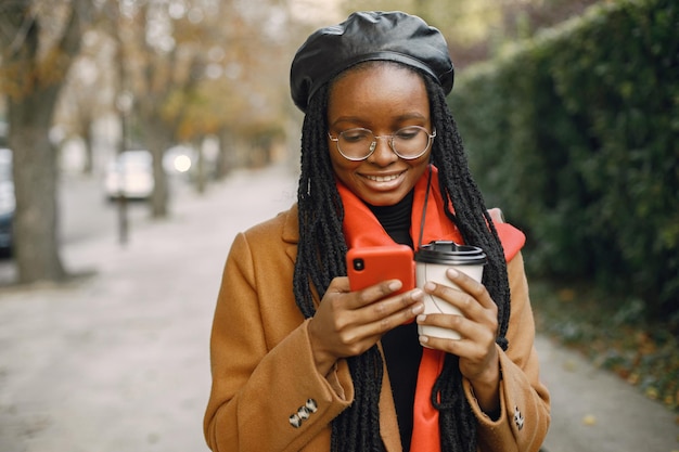 Joven mujer negra con un largo peinado de locomotoras de pie afuera con una taza de café para llevar. Mujer vestida con abrigo marrón, bufanda naranja y sombrero negro