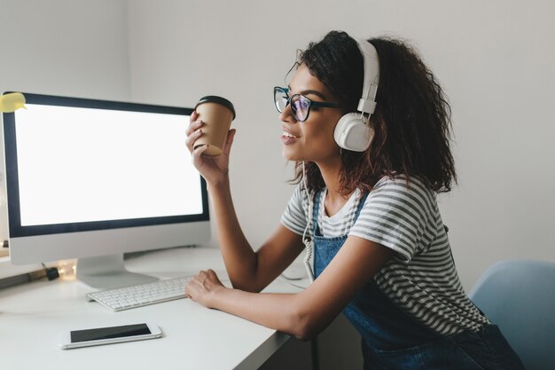 Joven mujer negra con nostalgia mirando a otro lado sosteniendo una taza de café y sonriendo mientras trabaja en la oficina