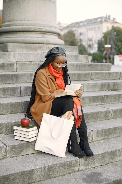 Joven mujer negra con peinados largos locs sentado en una escalera con un libro. Mujer vestida con abrigo marrón, bufanda naranja y sombrero negro