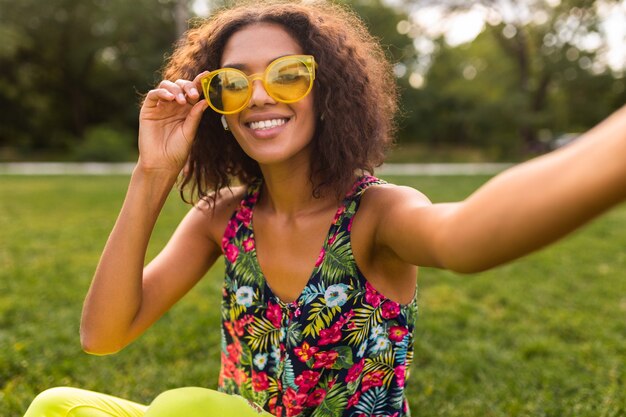 Joven mujer negra positiva con estilo tomando foto selfie escuchando música con auriculares inalámbricos divirtiéndose en el parque, estilo de moda de verano, traje colorido hipster