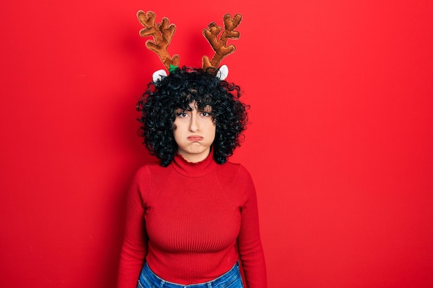 Joven mujer de oriente medio con lindos cuernos de reno de navidad hinchando las mejillas con cara graciosa. boca inflada de aire, expresión loca.