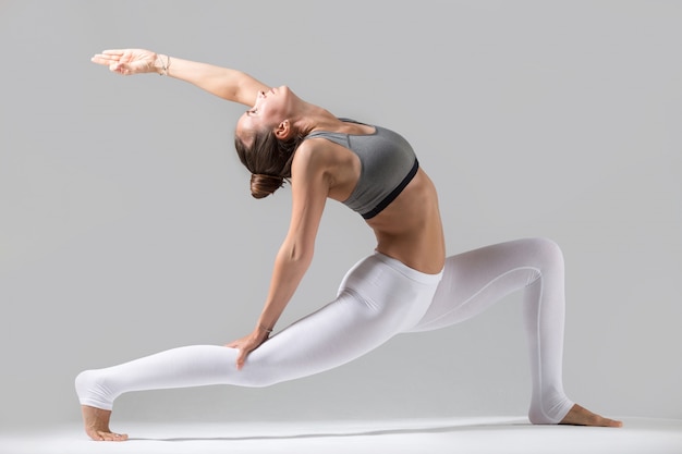 Joven mujer en pose anjaneyasana, fondo de estudio gris