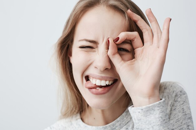 joven mujer positiva con cabello largo y rubio mirando a través de un gesto bien, mordiéndose la lengua con entusiasmo. Hermosa mujer rubia con ojos oscuros tiene buen humor juguetón.