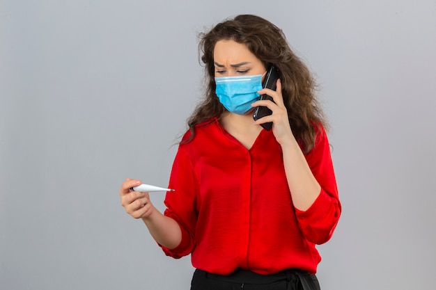 Joven mujer preocupada vistiendo blusa roja en máscara protectora médica hablando por teléfono móvil mirando termómetro digital sobre fondo blanco aislado