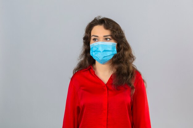 Joven mujer preocupada vistiendo blusa roja en máscara protectora médica mirando a otro lado sobre fondo blanco aislado