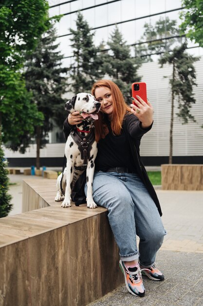 Joven mujer rojiza en la calle con su perro dálmata tomando una foto con el teléfono móvil